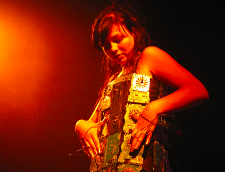 Meisje draagt jurk en bespeelt deze als muziekinstrument. Jurk bestaat uit allerlei verschillende plaatjes. Er komt een rode gloed van linksboven.